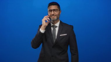 Mavi stüdyoda telefonu kulaktan kulağa tutan adam gülümserken takım elbise ve gözlük takıyor..