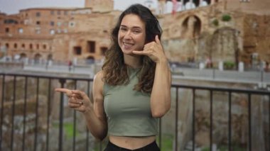 Latin kökenli genç bir kadın açık havada el hareketleriyle antik Roma harabeleri ile bağlantı kuruyor..