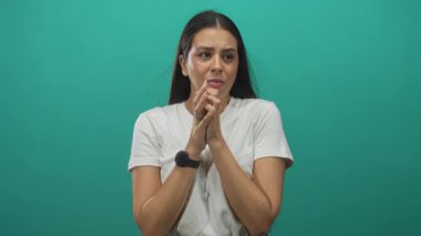 Göğüs tutan ve elini kenarda tutan bir kadın, beyaz bir t-shirt giyip yeşil bir stüdyoda seyrediyor, elleri göğsünde göğüs ağrısına işaret eden eller;.