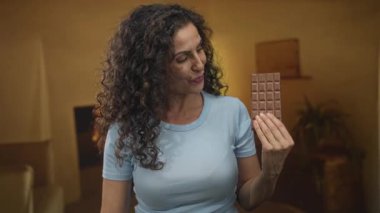 Koyu kıvırcık saçlı bir kadın stüdyo ortamında elinde çikolata tutarken gülümsüyor..