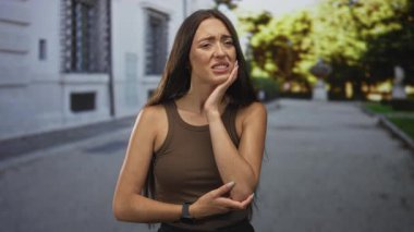 Güneş ışığıyla aydınlanan caddede elleri yanaklı bir kadın kolsuz tişört giyerken diş ağrısı ve endişeli somurtkanlık gösteriyor..