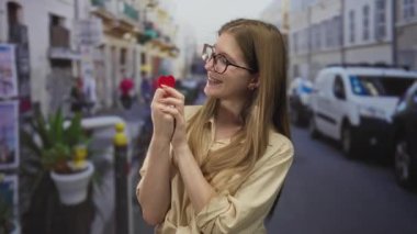 Yoğun bir sokakta küçük kırmızı bir kalbi tutarken gözlük takan ve gülümseyen genç sarışın kadın..