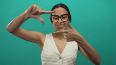 İzole edilmiş yeşil arka plan duvarına yaslanmış çerçeveli gözlüklü kadın güvenle gençlik yaratıcılığını ve ifadesini yakalıyor.