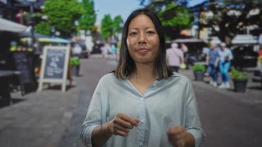 Kadın, açık mavi gömlek ve bilezik giyerek güneşli bir sokak pazarında kamerayı işaret ediyor. Gülümserken avuç içleri görünür..