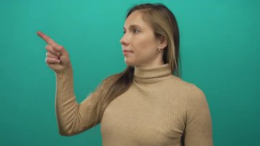 Balıkçı yaka kazaklı genç bir kadın, canlı yeşil bir duvarı işaret ederek, stüdyo ortamında izole edilmiş bir haldeyken kendine güvenen bir ifadeyle odak ve yönü aktarıyor..