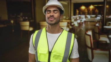 Şapkalı ve yansıtıcı yelek giyen genç mühendis restoranda barış işareti gösteriyor; zafer özgüveni.