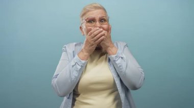 Kadın iki eliyle ağzını kapatır ve stüdyoda açık mavi bir arka plana bakar gözlük takar, kısa gri saçlar ve açık sarı bir üst; sürpriz.
