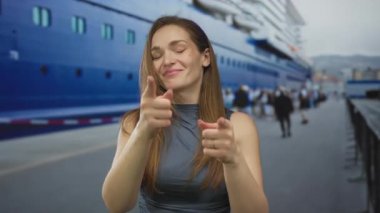 Çıplak kollu bir kadın, yoğun liman caddesinde, açık gökyüzü altında beyaz bir yolcu gemisinin yanında kameraya işaret ediyor..