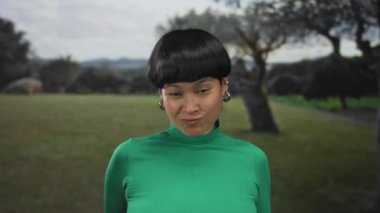 Yeşil elbiseli genç bir kadın dışarıda, yeşil bir parkta, neşeli bir ifadeyle rock 'n roll hareketi yapıyor..