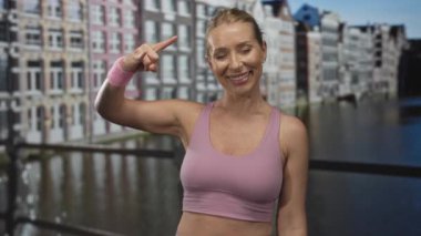 Pembe spor sutyen giyen kadın kaya boynuzu hareketi yapıyor ve Kanal Caddesi 'nde şehir binalarıyla gülüyor; eğlence enerjisi.