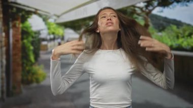 Uzun saçlı ve elleri havada bir kadın, yüzü caddeye bakıyor açık kafenin ve yürüyüş yolunun yanında el kol hareketi yapıyor; hayal kırıklığı.
