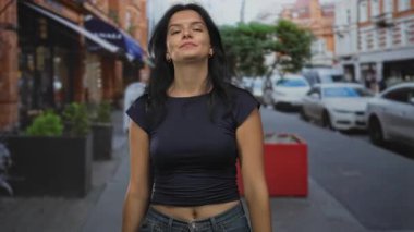 Yoğun caddelerde siyah gömlekli kadın kameraya kendinden emin bir gülümsemeyle işaret ediyor; davet.