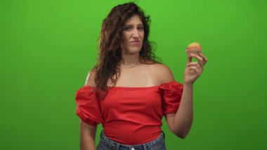 Kırmızı omuzlu bir kadın, elini kaldırmış kek tutuyor ve stüdyoda yeşil arka planlı tiksinmiş bir yüz; tiksinti, tiksinti, isteksizlik, tereddüt..
