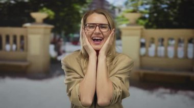 Şehir caddesinde şaşırmış bir kadın, sarı saçlı, gözlüklü, yanaklarındaki ellerle neşeyle gülümseyen, güneşli bir şehir zemininde yakalanan, mutluluk ve heyecan taşıyan bir kadın..