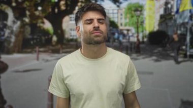 Bej gömlekli adam ağaçlar ve binalar tarafından çerçevelenmiş güneşli şehir caddesinde kameraya bakıyor..