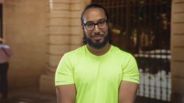 Sakallı ve gözlüklü adam bina girişinde görünür kolları olan neon yeşil gömlekli gülümsüyor; dostça bir nezaket..