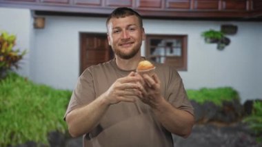 Adam gülümsüyor, iki eliyle kek tutuyor ve dışarıdaki bir binanın girişinde kameraya işaret ediyor; neşeli bir karşılama..