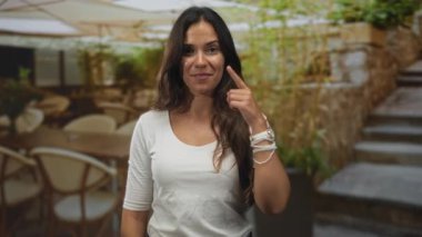 Kadın, bileklik ve bilekliklerle gülümseyen, güneşli restoran terasında parmak yanağa işaret ediyor; oyuncu bir güven.
