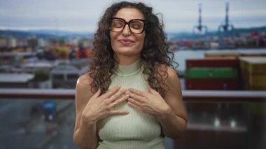 Kadın, arkasında nakliye konteynırları ve vinçlerle birlikte deniz kıyısındaki bir rıhtımda göğüs göğüse bir balıkçı yaka kazak giyiyor..