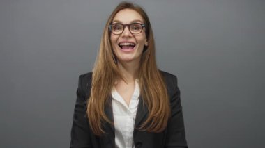 Gözlüklü, siyah ceketli, beyaz tişörtlü, ağzı açık gri stüdyo arka planına karşı konuşan bir kadın..