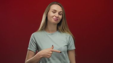 Uzun saçlı bir kadın izole edilmiş kırmızı bir arkaplanı işaret ederek gülümsüyor. Kendine güvenini gösteriyor ve arkadaşça bir tavır sergiliyor.