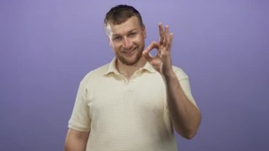 Sağ eliyle OK işareti gösteren ve mor bir stüdyo ortamında gülümseyen bir adam krem rengi polo tişört giyiyor; güven onayı.