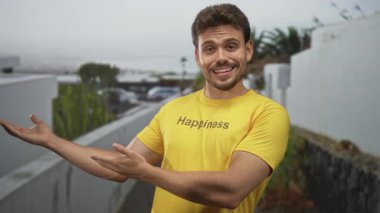 Elleri kalçasında poz veren, sarı tişörtlü, baskılı mutluluk giyen, beyaz bir duvarın yanında hafifçe sırıtan bir adam ve sokakta kaktüs; mutluluk..