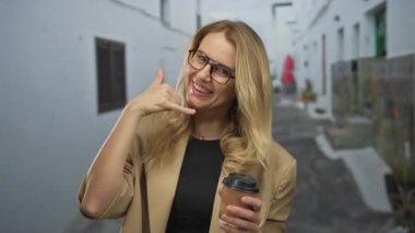 Elinde kahve fincanı ile gülümseyen bir kadın gündüz vakti şehir sokaklarında gözlüklü ve bej ceketli bir şekilde telefon jesti yapıyor.