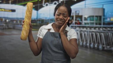 Önlüklü kadın fırıncı bir eliyle uzun bir baget tutuyor ve havaalanı terminalinde bavul vagonları ve tabelalarıyla diş ağrısı için parmaklarıyla yanağına bastırıyor..
