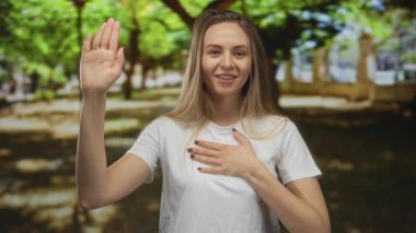 Genç bir kadın, canlı yeşil ağaçlarla çevrili güneşli bir parkta yemin ediyor, samimi bir ifadeyle, elini kaldırıyor ve kalbini tutuyor, bir anlık sadakati somutlaştırıyor.