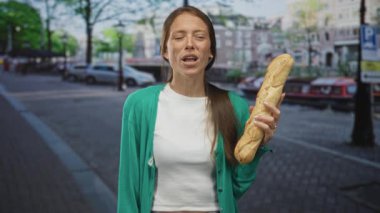 Şehir caddesinde elinde baget tutan bir kadın yeşil hırka ve beyaz hasat bluzu giyiyor, gözleri kapalı ve rahat bir şekilde gülümsüyor; neşe.