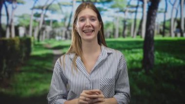 Sarışın kadın çizgili gömlek giyiyor, el çırpıyor, ormanda geniş gülüşlü, açık yeşil ağaçlı, heyecan dolu..