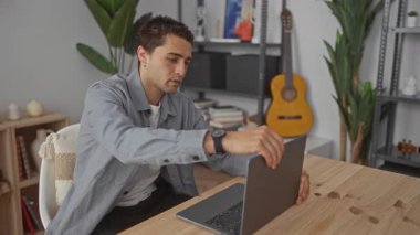 Genç İspanyol bir adam rahat oturma odasında dizüstü bilgisayarda çalışıyor. Arka planda gitar var. Yaratıcılık ve odaklanma konusunda rahat bir iç atmosfer sergiliyor..