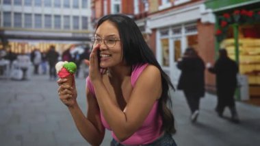 Meşgul bir caddede dükkan önleri ve yayalarla konuşurken elinde üç kepçe dondurma külahı tutan ve kıt kanaat ağza alan kadın, eğlenceli bir merak..