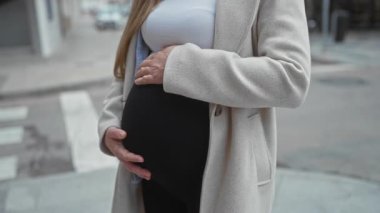 Hamile kadın şehir caddesinde dışarıda duruyor, palto giyiyor, karnını tutuyor, modern bir ortamda anneliğin güzelliğini ve beklentisini gösteriyor..