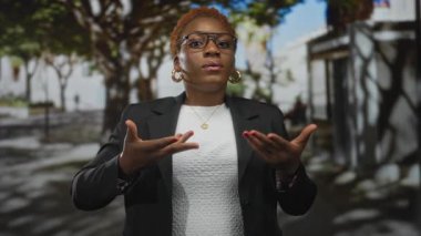 Gözlüklü kadın sokaklarda yeşil ağaçlarla işaret yapıyor. Güneş ışığı gölgeleri yansıtıyor..