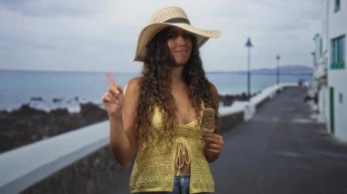 Saman güneş şapkası takan İspanyol genç kadın çikolatalı fındıklı dondurma tutuyor ve bir deniz kenarı caddesini işaret ediyor..