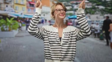 İşaret parmağıyla işaret eden ve gülümseyen, gözlüklü ve çizgili hırka giyen bir kadın, kaldırım taşı bir kafede şemsiyeli..