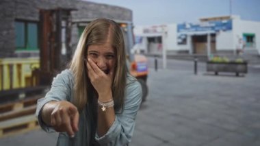 Sarışın genç bir kadın, şehir caddesinde ağzı kahkahalarla doldururken kamerayı işaret ediyor..