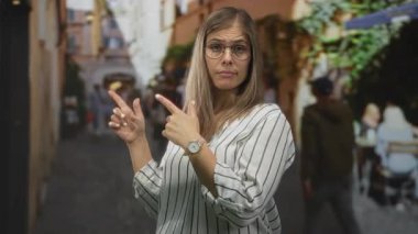 Gözlüklü sarışın kadın dışarıdaki kafe masalarının yanındaki dar kaldırım taşı sokağını işaret ediyor; belirsizlik..