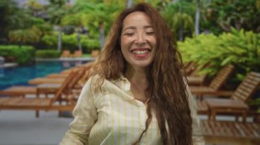 Gülümseyen kadın, otel havuzunda kollarını kaldırıyor çizgili tişört giyiyor açık havada güneşli yataklar ve palmiyeli tatil beldesinde yüzünü ve ellerini gösteriyor; keyif tatili kutlaması.