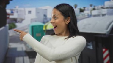 İspanyol kadın, güneşli bir sokakta, geri dönüşüm kutularının yanında, kentsel bir ortamda olumlu çevresel eylemlerin altını çizerek mutlu bir şekilde baş parmak gösteriyor..