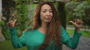 Ormanda göğsüne işaret eden başparmaklı kadın yeşil kazak ve uzun saç giyiyor, hafif ekşitme; umursamazlık.