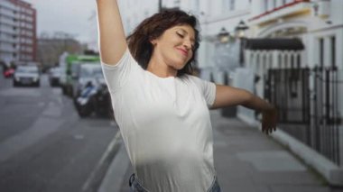 Kadın kolunu kaldırır, çilli yüzü güler, beyaz tişört giyer ve kot pantolon giyer; kaygısızca eğlenir..