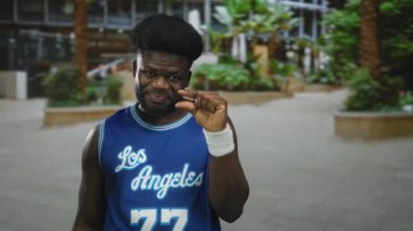 Bir bina meydanının önünde parmaklarını çimdikleyen adam mavi basketbol forması giyiyor..