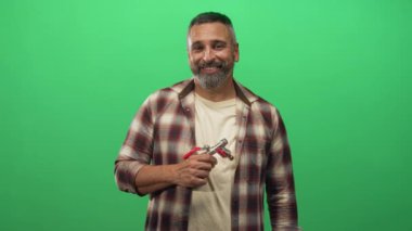 Orta yaşlı bir adam elinde musluk aleti tutarak yeşil stüdyo setindeki gülüşünü işaret ediyor..