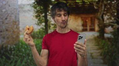 Adam binanın önünde sırıtırken yarısı yenmiş donut ve akıllı telefon tutuyor..