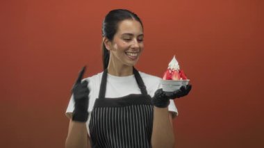 Turuncu stüdyoda siyah eldiven giyerken çilekli tatlı kasesini gösteren önlüklü genç esmer kadın; neşeli bir konukseverlik..