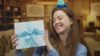 Mavi kurdeleli bir hediye kutusu tutan ve rahat oturma odasında gülümserken parti şapkası takan bir kadın..