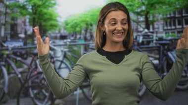 Elleri havada gülümseyen, yeşil bir kazak giyen, Amsterdam 'da park etmiş bisikletli bir kanalın kenarında oturan bir kadın..